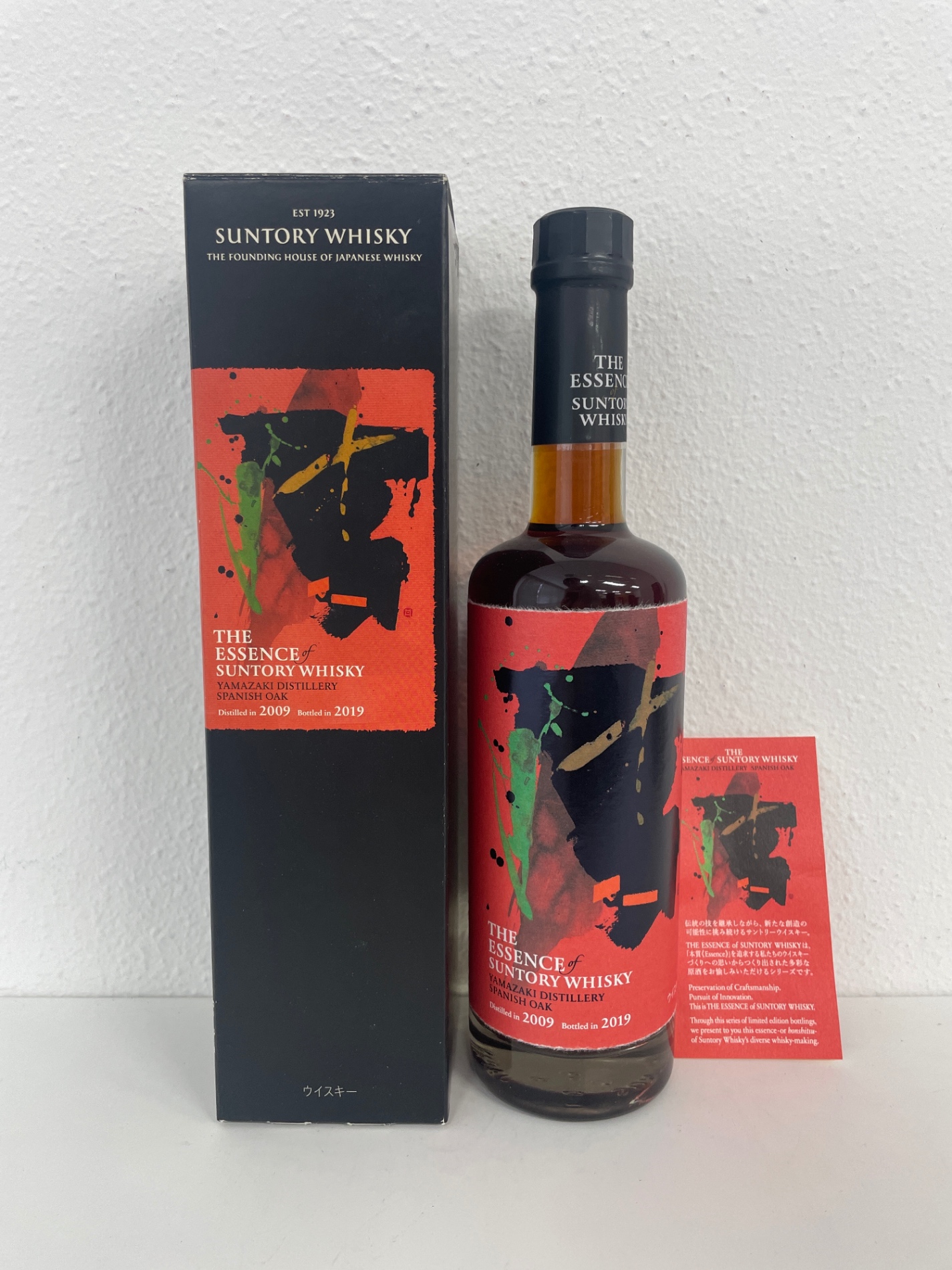 Bottle of The Essence of Suntory Whisky with a red abstract label standing beside its black box on a white surface