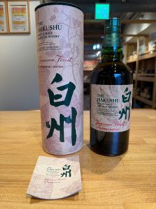 Bottle of The Hakushu Japanese Whisky with a pink forest pattern label and a matching cylindrical box on a wooden table Bittersweet Edition Japanese Forest