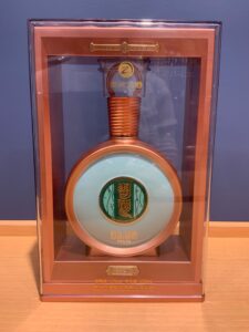Copper toned commemorative liquor bottle on display in a clear case with a green circular label and Chinese characters visible on the seal piece above the stem area