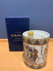 Decorative ceramic cognac tin with a lid and Napoleon era scene next to a dark blue CAMUS box on a wooden table