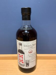 Whiskey bottle with a cream label reading Ichiros Malt Card on a wooden surface against a blue wall