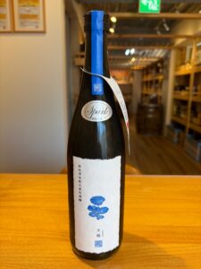 Tall sake bottle on a wooden counter with a blue tape seal on the neck and a white label featuring a blue floral design store shelves blurred in the background