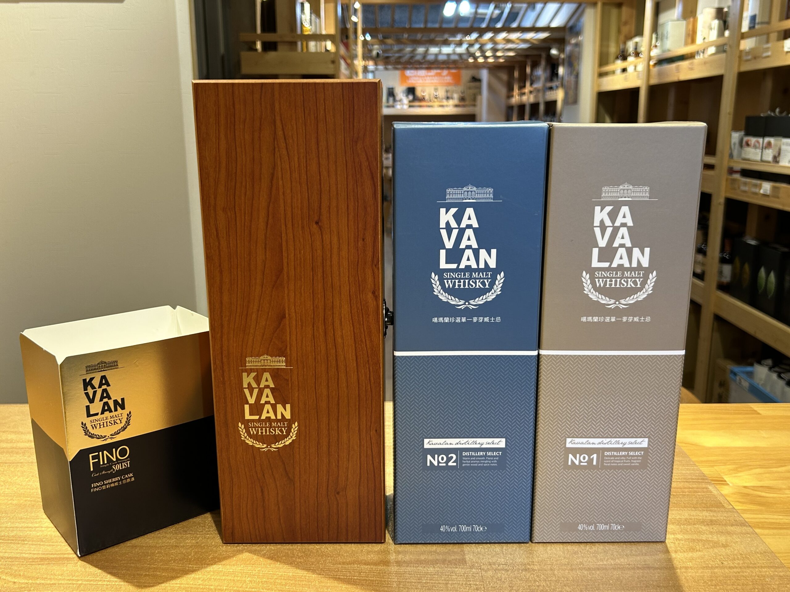 Row of Kavalan whisky gift boxes in blue and gray standing on a wooden shelf with a wood grain panel in front and a store background behind