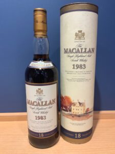 Macallan 18 Years Old single malt Scotch whisky bottle beside its tall cylindrical box against a blue wall