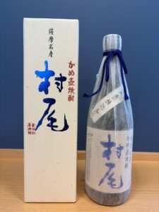 Box and wrapped bottle of Japanese sake or shochu with blue calligraphic characters and red stamp, on a wooden surface against a dark background.