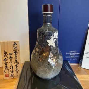 Decorative dark glass bottle with floral pattern on a wooden surface beside blue display boxes and a wooden Japanese inscription plaque.