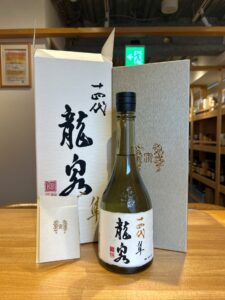 Bottle of sake with a white label and elegant black kanji flanked by white and silver gift boxes on a wooden counter in a shop setting