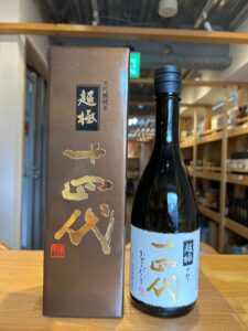 Dark glass sake bottle with a light blue label and gold Japanese characters placed beside a tall brown box on a wooden table in a shop installation