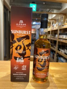Bottle of Sunburst World Malt whisky with orange label beside its tall red box both labeled Nagahama Distillery Japan and Edition KIWAMI on a wooden counter in a store