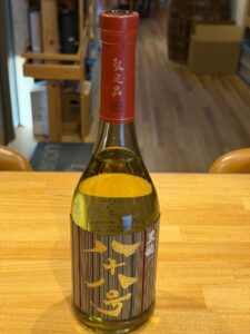 Close up of a bottle on a wooden table red foil neck with vertical gold Japanese characters and a decorative striped label on the body of the bottle background of a shop