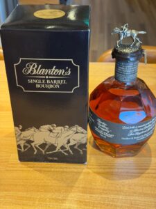 Bottle of Blantons Single Barrel Bourbon next to its black box on a wooden surface 750 ml label visible on the box and bottle cap featuring a horse stopper that resembles a horse statue across the top of the bottle neck