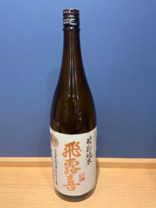 Brown glass bottle of sake with a white label showing large orange Japanese characters placed on a wooden table against a blue wall