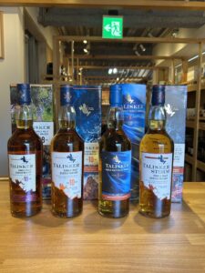 1 横浜と東京のお酒買取専門店LIQUORJOYリカージョイ Row of Talisker single malt whisky bottles with blue and white boxes on a wooden display in a store window