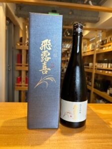 1 お酒買取専門店LIQUORJOYリカージョイ Japanese sake bottle beside a dark blue gift box with gold Japanese characters and a decorative grain motif on a wooden counter