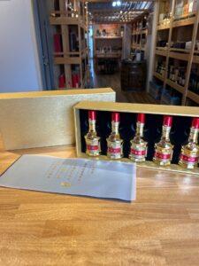 1 お酒買取専門店LIQUORJOYリカージョイ Row of gold labeled liquor bottles in a wooden display case on a shop counter with shelves and a tasting area in the background