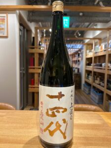 1 お酒買取専門店LIQUORJOYリカージョイ Tall dark bottle of sake with a beige label featuring large brown kanji placed on a wooden table in a shop setting