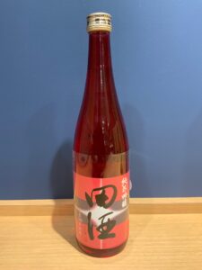 Tall glass bottle of red liquid with a pink Japanese label capped with Japanese text on a wooden surface against a blue wall