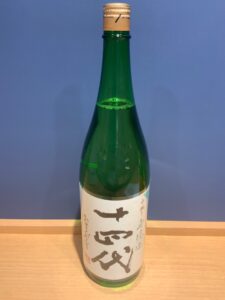4atlRxfhTsQSvnN0JdNZqmEC1CFtRZhTf5CaVKOG お酒買取専門店LIQUORJOYリカージョイ Green sake bottle with a white label and large black kanji characters on a wooden counter against a blue wall