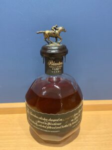 Bottle of Blantons Single Barrel Bourbon with a brass horse and jockey stopper on top of the neck against a blue wall background