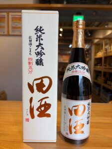 White box with bold black Japanese characters stands beside a dark sake bottle wrapped in gold foil in a store setting.