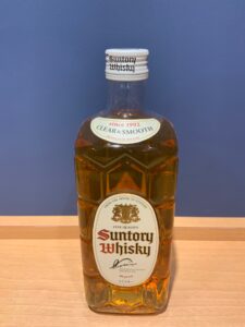 6tyTCHb64mFHUBbpYoUbqzCyqwjd2gDcp4G4dIwU お酒買取専門店LIQUORJOYリカージョイ Bottle of Suntory Whisky with a white cap and amber liquid label reads Suntory Whisky on a wooden surface against a blue wall
