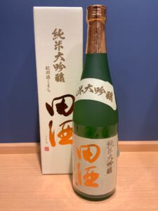 Green glass sake bottle with foil wrapped neck beside a white box with gold Japanese characters label shows orange kanji text