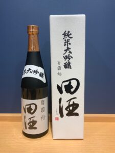 Japanese sake bottle with copper foil cap beside a white box with black Japanese calligraphy on a wooden surface