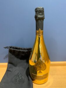 acUGSfyWGYC747IBmUpHBaLcdYN0VxVTbXZTrnOh 横浜と東京のお酒買取専門店LIQUORJOYリカージョイ Champagne bottle with black foil at the neck and gold emblem resting on a wooden surface beside a black fabric pouch