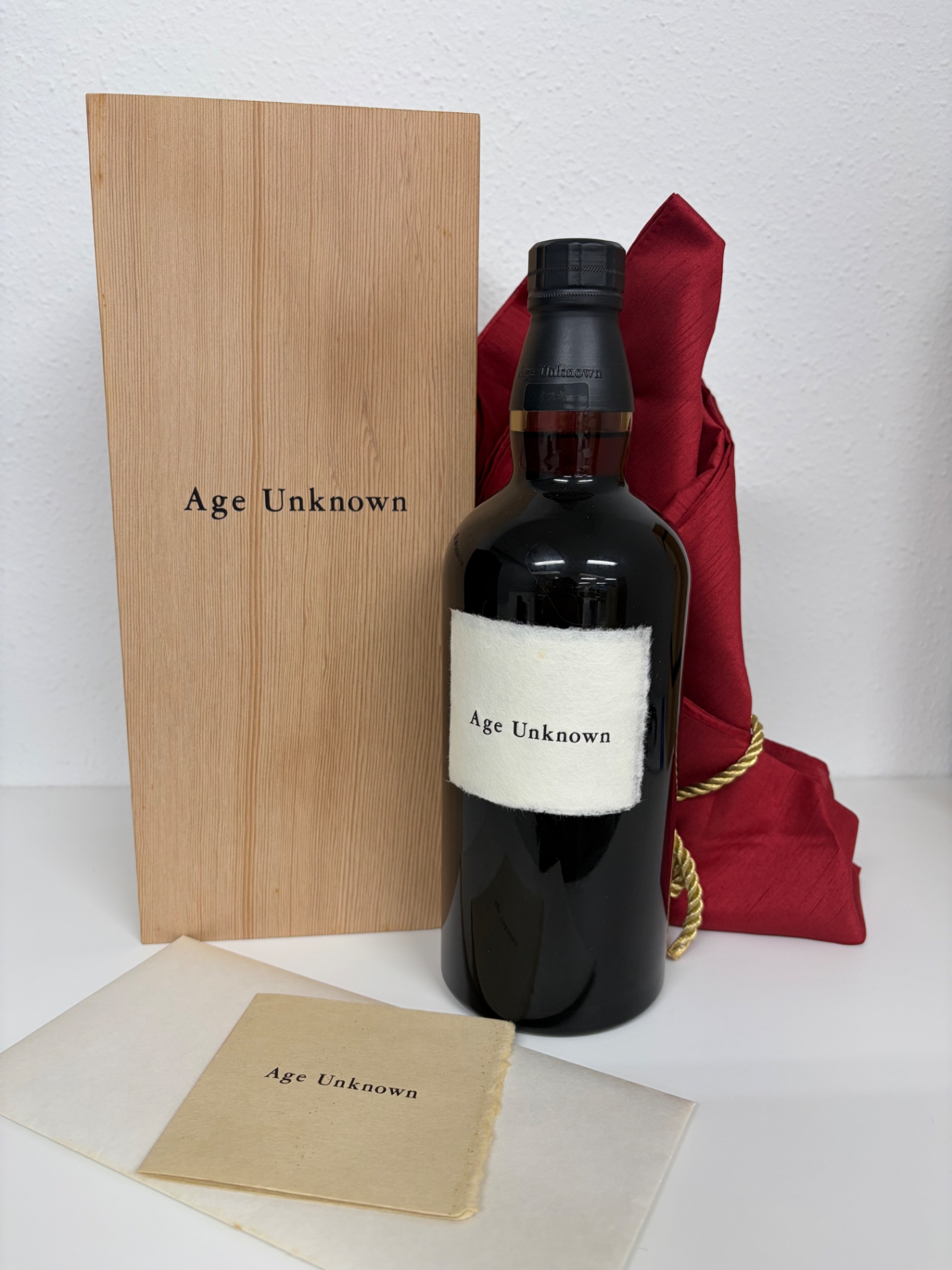 Wine bottle with a light colored label placed beside a wooden gift box that reads Age Unknown and a red cloth in the background on a white surface