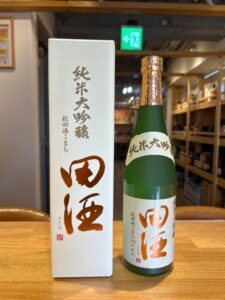 Green bottle of sake with copper foil cap next to a white gift box with vertical Japanese text, on a wooden counter with store shelves in the background.