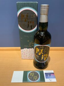 Bottle of Akkeshi Japanese whisky beside its decorative green box on a wooden surface with a folded brochure in front