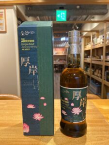 Bottle of The Akkeshi Single Malt Japanese Whisky (Peated) beside its blue-green decorative box with pink lotus motifs on a wooden table.