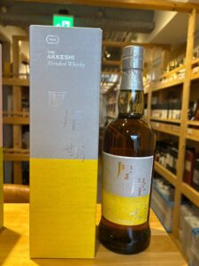 Bottle of The Akkeshi Blended Whisky beside its gray–yellow boxed packaging on a wooden counter in a store.