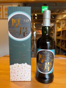 Box and bottle of Akkeshi The Akkeshi Single Malt Japanese Whisky on a wooden table, blue packaging with gold Japanese characters and pink cherry blossoms at the bottom.