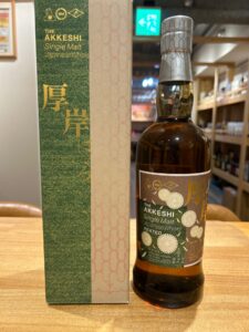 Bottle of The Akkeshi Single Malt Japanese Whisky next to its green and beige patterned box on a wooden surface.