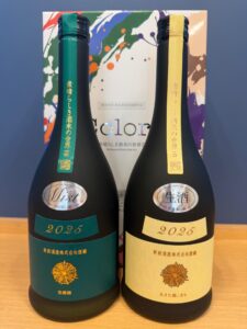 Two dark sake bottles with teal and gold vertical labels reading 2025 standing on a wooden surface with a colorful abstract box in the background