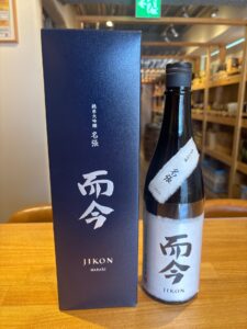 Dark sake bottle with a white paper label next to a navy box with white Japanese characters on a wooden counter in a shop setting