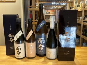 Row of sake bottles with dark labels and boxed packaging on a wooden counter inside a shop