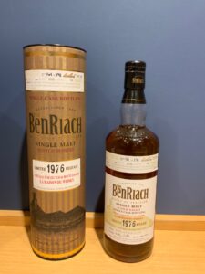 Two BenRiach single malt Scotch whisky bottles standing side by side against a blue wall one in a tall brown gift tin labeled 1976 release on the left and a bottle with a black cap on the right