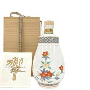 White ceramic bottle with colorful floral design and wooden cap beside a wooden sake box and a cream booklet
