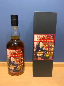 Whiskey bottle labeled Ichiros Malt Chichibu beside its black gift box on a wooden surface against a blue wall