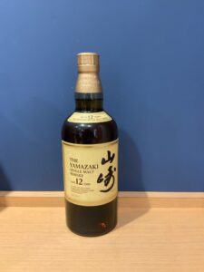 LGDfFaOH3urNYMEMjFnWqe8DC0HcPqhBrA6lW6nY お酒買取専門店LIQUORJOYリカージョイ Bottle of The Yamazaki 12 year old single malt whisky on a light wooden surface against a blue wall label facing forward