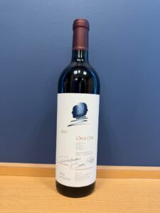Bottle of Opus One 2019 red wine standing on a wooden surface against a blue wall label with blue logo and signatures visible