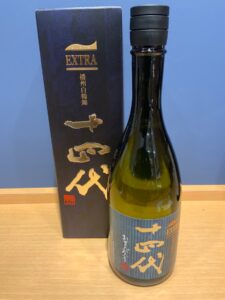 abtHT1Q93L9EA9O4d12ETMgGwnHy0lvUKXKS3tYg お酒買取専門店LIQUORJOYリカージョイ Sake bottle with dark glass and a blue striped label standing beside a tall navy box with gold EXTRA lettering and Japanese characters