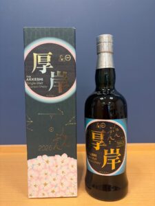 Bottle of Akkeshi Single Malt Japanese Whisky beside its decorative box with pink blossoms and metallic cap detail on a wooden surface on a blue background