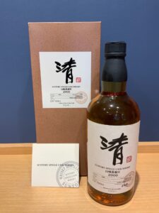 Whiskey bottle with white label beside a brown box against a blue wall label shows Japanese kanji and Suntory Single Cask Whisky 2000 wording