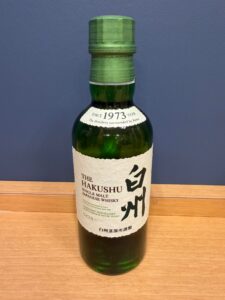 Bottle of The Hakushu single malt Japanese whisky with beige label green glass and green cap on a wooden surface against a blue wall