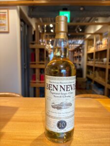 A bottle of Ben Nevis Highland Single Malt Scotch Whisky on a wooden table with a shop background and shelves behind it