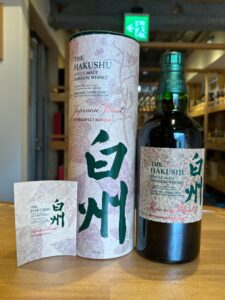 The Hakushu Japanese Forest Bittersweet Edition whisky bottle beside a textured beige box and matching labels on a wooden counter with a small card in front