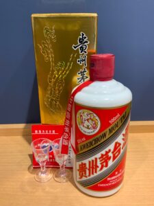 Large white bottle of Kweichow Moutai with red cap and red label placed beside a tall gold decorative box red ribbon draped over the bottle on a wooden table with small clear shot glasses nearby